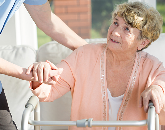 Female nursing home patient with a walker being assisted to stand by a staff member who is out of frame.