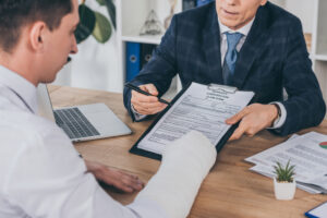 businessman in blue jacket giving form for compensation claim to worker with broken arm at table in office