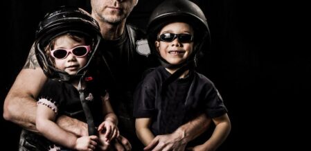 Image of a father with two young children smiling and wearing motorcycle helmets and goggles