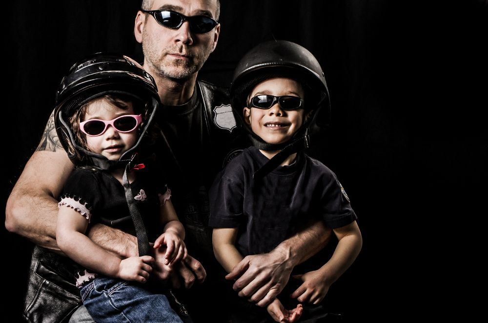 Image of a father with two young children smiling and wearing motorcycle helmets and goggles