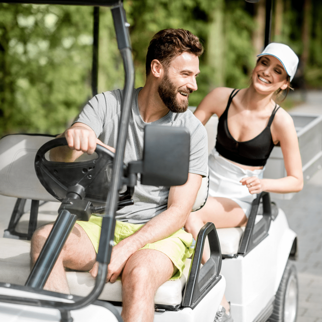 Man driving a golf cart with one hand on the wheel, looking over his shoulder and talking with woman in the backseat.