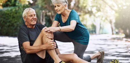 An elderly man is on the ground having knee pain while an elderly woman is trying to help him