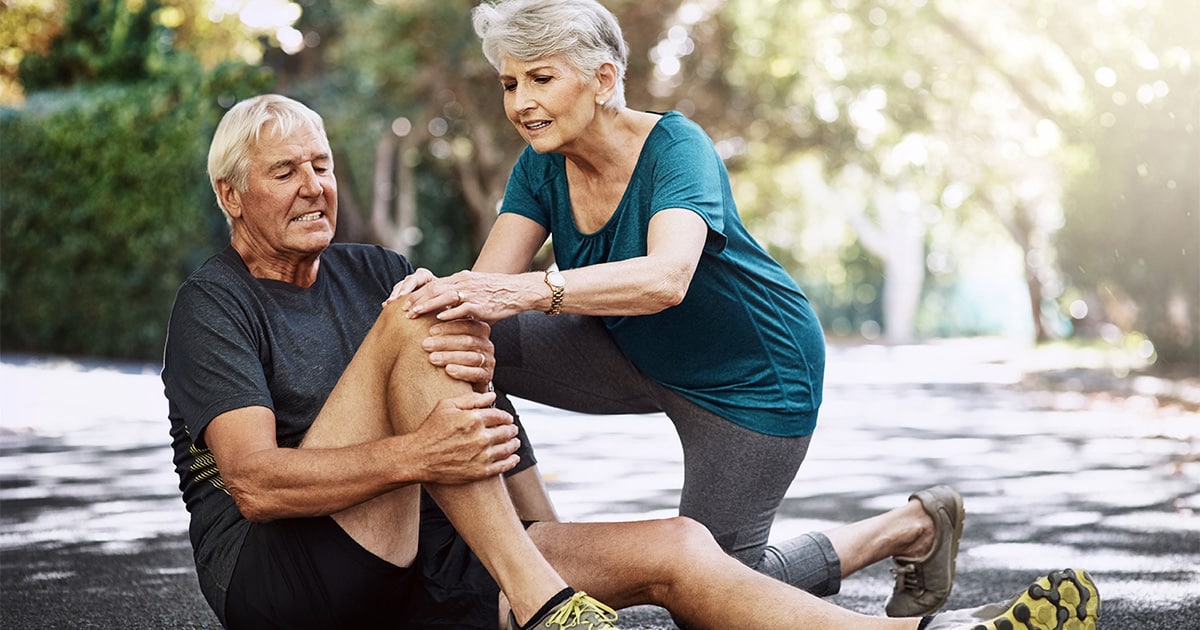 An elderly man is on the ground having knee pain while an elderly woman is trying to help him