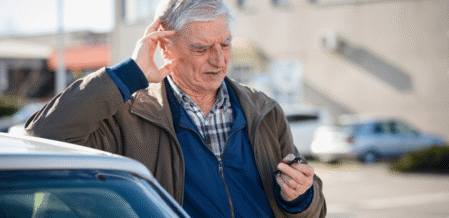 Image of a man looking confused and in pain with a hand over his ear, and leaning against a car