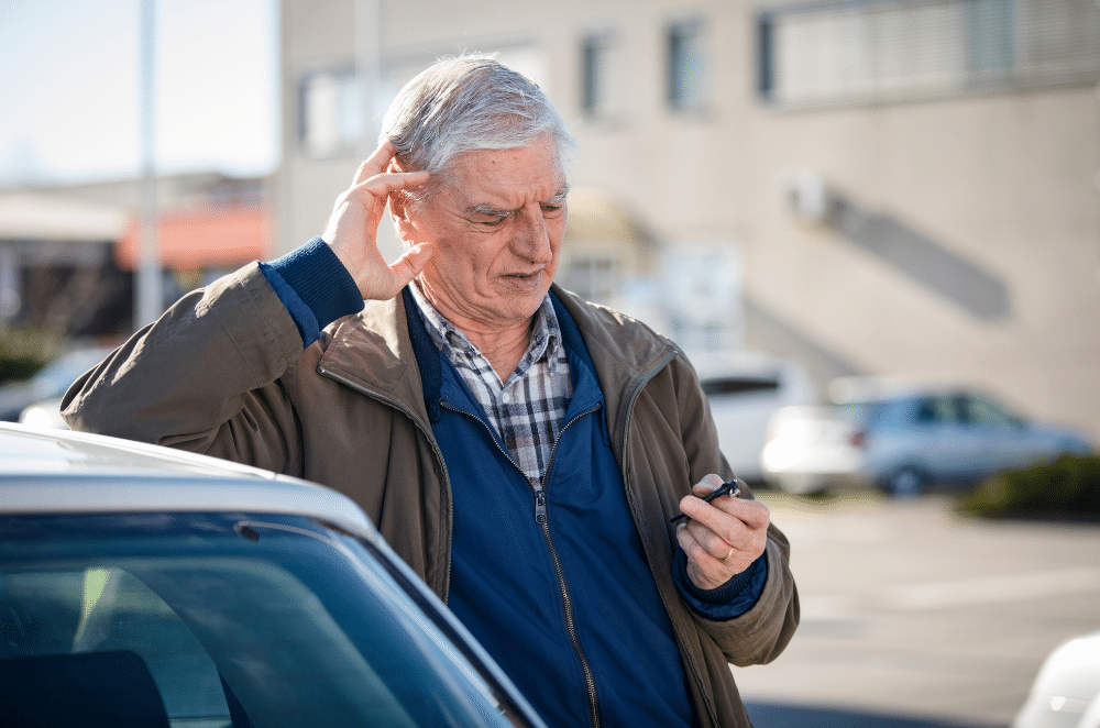 Image of a man looking confused and in pain with a hand over his ear, and leaning against a car