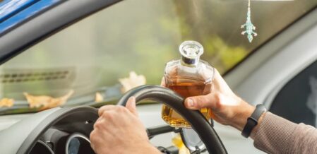 The driver sits behind the wheel, holding an alcoholic beverage, breaking the law by drinking while driving.