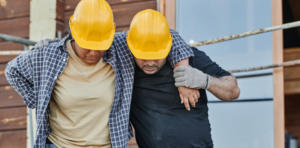 Construction worker helps injured colleague walk off-site, both wearing yellow hard hats and plaid shirts